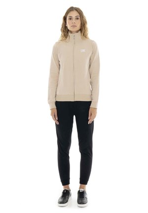 Une femme porte une veste beige zippée, un pantalon de jogging noir et des baskets noires sur fond blanc.