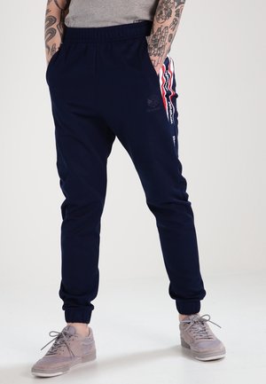 Persona con joggers azul marino con franjas laterales rojas y blancas, zapatillas grises y camiseta gris, de pie con las manos en los bolsillos.