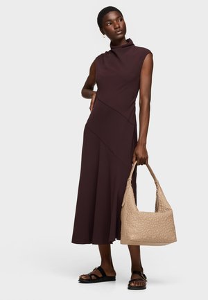 Donna che indossa un vestito midi senza maniche marrone scuro e sandali neri, con in mano destra una borsa beige texturizzata.