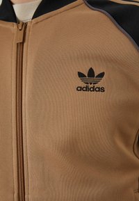 Ruskea vetoketjullinen takki, joka on valmistettu teksturoidusta kankaasta ja jossa on musta Adidas-logo sekä kontrastiväriset mustat yksityiskohdat kauluksessa ja olkapäillä.