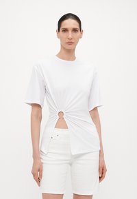 Hvit bomull T-skjorte med korte ermer, med en vridd design og sirkulært åpning i midjen. Brukt med hvite shorts.