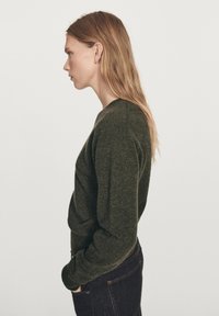 Femme aux longs cheveux blonds portant un pull à manches longues vert foncé et un jean foncé, debout de profil les mains dans les poches, sur un fond uni.