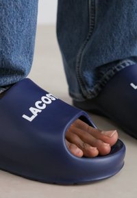 Sandales en caoutchouc bleu marine avec un bout ouvert et un grand logo blanc "LACOSTE" sur le dessus. Semelle intérieure texturée et semelle lisse.