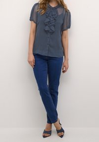 Blå volangblus med korta genomskinliga ärmar, texturerad tyg, knäppning, parat med blå denimjeans och mörkblå klacksandaler.