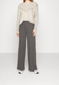 Jasnobeżowy sweter z koronkową lamówką i falbankami przy dekolcie, zestawiony z szarymi, żeberkowymi spodniami o szerokich nogawkach, noszony z białymi tenisówkami.