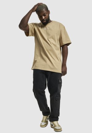 Hombre con cabello rubio corto, vestido con una camiseta beige de gran tamaño, pantalones cargo negros y zapatillas beige, de pie con una mano sobre la cabeza.