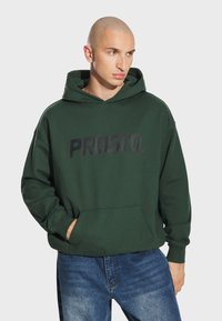 Zielona bluza z kapturem z czarnym logo "PROSTO." na piersi, z przednią kieszenią i ściągaczami przy mankietach. Połączona z niebieskimi dżinsami.