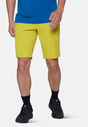 Mann trägt gelbe Shorts, schwarze Socken, schwarze Sportschuhe und ein blaues Hemd und steht vor einem einfarbigen hellen Hintergrund.