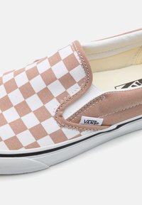 Chaussure Slip-on Vans avec motif damier beige et blanc, accents élastiques sur les côtés, semelle blanche avec bande noire et étiquette de marque visible.