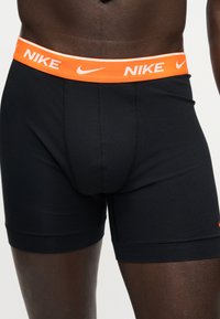 Άνδρας που φοράει μαύρα μπόξερ Nike με πορτοκαλί μέση που φέρει λευκά λογότυπα Nike και σύμβολα swoosh.