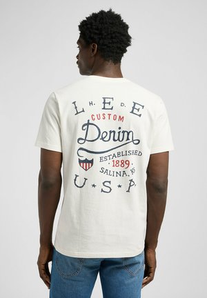 T-shirt color crema con scollatura rotonda, caratterizzato da testo stampato blu e rosso sul retro, fra cui "LEE CUSTOM Denim" e un logo a scudo.