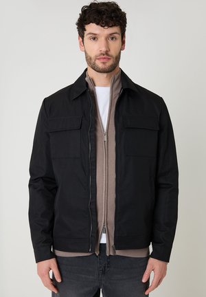 Mężczyzna o ciemnych, kręconych włosach, ubrany w czarną kurtkę na beżowym swetrze zapinanym na zamek i białą koszulkę, stojący na jednolitym tle.