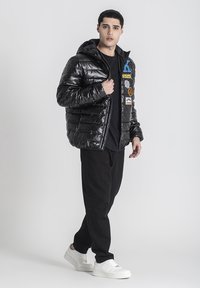 Chaqueta acolchada negra y brillante con capucha, que presenta secciones acolchadas y varios parches. Llevada sobre una camiseta negra y combinada con pantalones negros y zapatillas blancas.