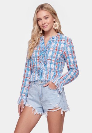 Blond kvinde iført store vævede ringearringe, en blå og rød ternet jakke med frynser og lyse slidte denimshorts med den ene hånd i lommen.