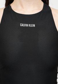 Κοντινό πλάνο ενός ατόμου που φοράει μαύρο υφασμάτινο αμάνικο τοπ με το λογότυπο "CALVIN KLEIN" τυπωμένο με λευκά γράμματα στο στήθος.