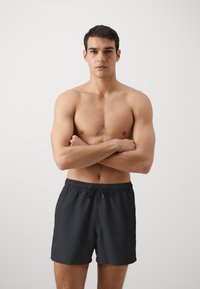 Crne kupaće hlače s elastičnim pojasom i vezicom. Glatka tkanina bez vidljivih uzoraka. Model stoji sa prekriženim rukama.
