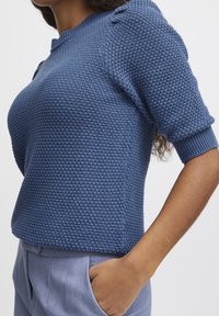 Modrý svetr s krátkými rukávy a texturovaným pleteným vzorem, žebrovanými manžetami a kulatým výstřihem, doplněný světle modrými kalhotami.