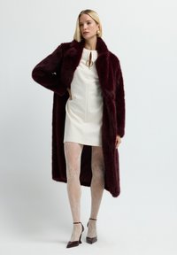 Nainen, jolla on päällään burgundinvärinen tekoturkki ja valkoinen mini mekko, kuviolliset läpinäkyvät sukkahousut ja burgundinväriset kärkitossut, poseeraa käsi lantiolla.