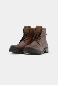 Botas de tobillo de cuero marrón con cordones, suelas de goma, cuellos acolchados y detalle de correa con hebilla sobre un fondo blanco.