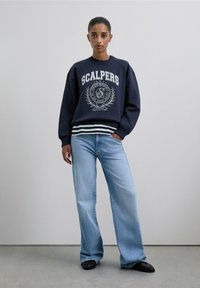 Sudadera navy con impresión blanca, que presenta "SCALPERS" y diseño de emblema. Combinada con jeans de pierna ancha azul claro y zapatos negros.