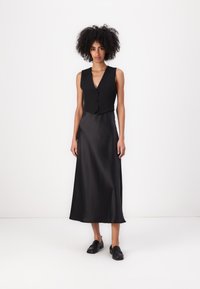 Zwarte vest met knopen, gecombineerd met een lange, vloeiende zwarte satijnen rok. Schoenen zijn zwarte instappers. De outfit heeft een gladde textuur.