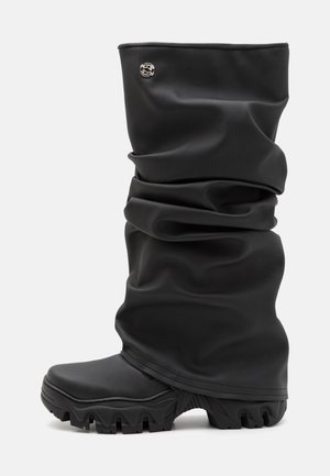 Botas de goma negras con diseño arrugado, parte superior texturizada, suela gruesa y detalle de hardware plateado en el lateral.