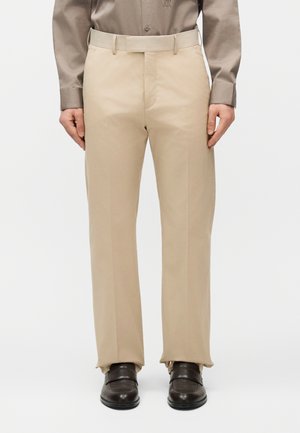 Mand iført beige habitbukser, mørkebrune lædersko og en langærmet taupefarvet skjorte med knappede manchetter.