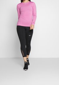 Růžový sportovní top s dlouhým rukávem spojený s černými legínami s průsvitnými detaily, oba vyrobené z pružné, odvádějící pot tkaniny. Černé tenisky.