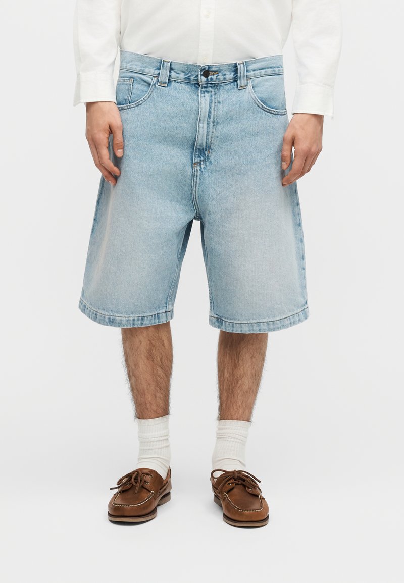 Homme portant un short en jean bleu clair, des chaussettes blanches, des chaussures bateau marron et une chemise blanche à manches longues, debout devant un fond blanc.