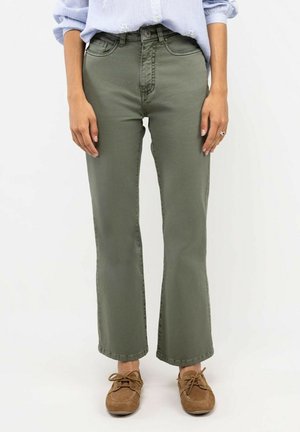 Persona con pantalones rectos verde oliva, camisa de manga larga azul claro remangada y zapatos marrones con cordones, de pie frente a un fondo blanco.
