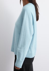 Vaaleansininen neulepaita pehmeästä kankaasta, jossa on rento leikkaus, ribsikimpput ja pyöreä pääntie. Malli esitetään sivusta.