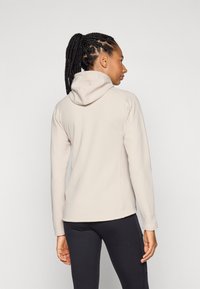 Veste beige claire à capuche avec un tissu texturé, manches longues et un design ajusté, vue de dos, associée à un legging noir.