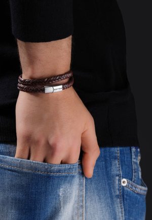 Bracelet en cuir tressé marron avec un fermoir en argent, porté au poignet. Présente un accent métallique plat ; main reposant dans une poche en denim.