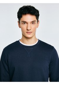 Młody mężczyzna o krótkich, kręconych czarnych włosach, ubrany w ciemnogranatową bluzę na białej koszulce z okrągłym dekoltem, patrzący przed siebie z neutralnym wyrazem twarzy.