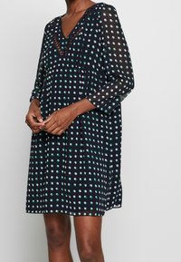 Kvinna i en mörkblå klänning med gröna och vita prickar, V-ringning med spetsdetaljer och trekvarts transparenta ärmar.