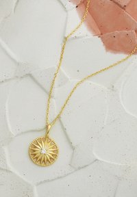 Guldhalskæde med en rund vedhæng med et solstråledesign og en lille klar ædelsten i midten, hvilende på en tekstureret overflade.