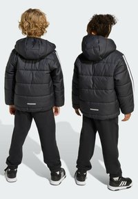 Vestes rembourrées noires avec capuches, présentant des rayures blanches le long des manches, portées avec des pantalons de survêtement noirs et des baskets noires. Détails réfléchissants inclus.