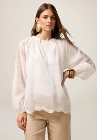 Blusa blanca con bordado floral, corte relajado, mangas abullonadas, dobladillo con forma de ondas y un escote con dos detalles de botones. Usada con pantalones beige.
