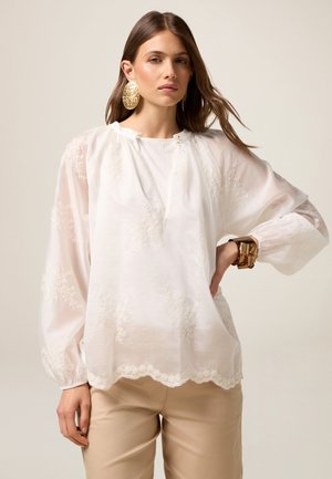 Camicia bianca con ricami floreali, vestibilità comoda, maniche a palloncino, orlo scallopato e scollatura con due dettagli a bottone. Indossata con pantaloni beige.