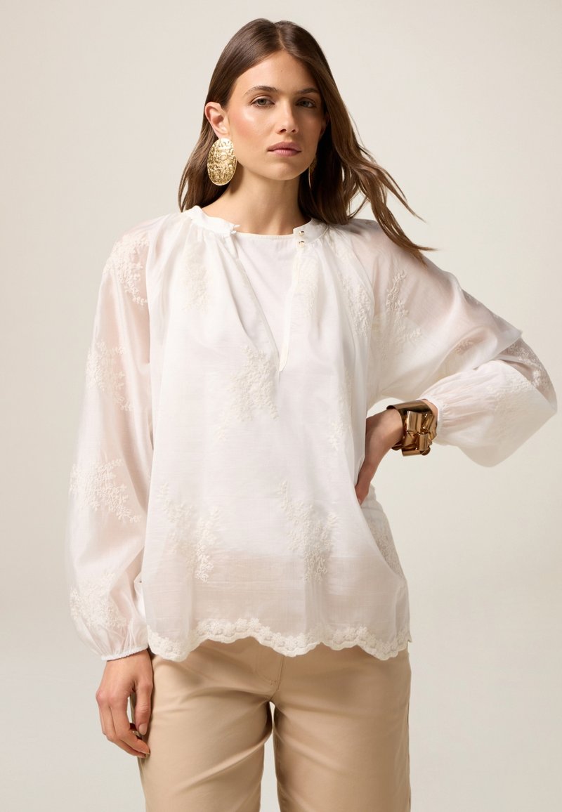 Blusa blanca con bordado floral, corte relajado, mangas abullonadas, dobladillo con forma de ondas y un escote con dos detalles de botones. Usada con pantalones beige.