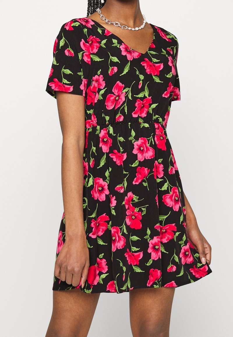 Robe noire à manches courtes avec motif floral rose vif, taille cintrée, portée par une personne aux cheveux tressés et un collier en argent et perles.