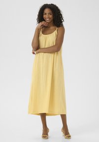 Gelbes ärmelloses Maxikleid mit gerafftem Ausschnitt, fließendem Stoff und knöchellanger Silhouette. Getragen mit gelben Absatzsandalen.