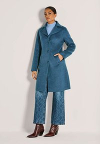 Vrouw in een tealblauw knielang jasje, met gemønstarde wijde jeans, bruine haklaarzen en een lichtblauwe coltrui.