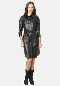 Vestido camisa de cuero negro con mangas largas, bolsillos en el pecho y cintura ceñida con cinturón. Presenta un cierre frontal de botones y una textura suave.