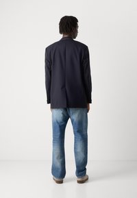 Blazer azul marinho com ombros estruturados e costura contrastante, combinado com jeans azul claro e sapatos cinzentos, visto de costas.