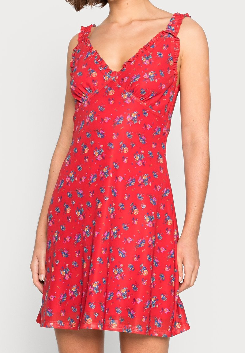 Robe rouge sans manches avec un petit motif floral multicolore, bretelles à volants et encolure en V portée par une personne debout.
