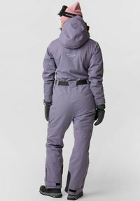 Paars waterdichte skikostuum met een figuurgetailleerd ontwerp, capuchon, zwarte accenten op de manchetten en een verstelbare tailleband. Gecombineerd met handschoenen en een roze muts.