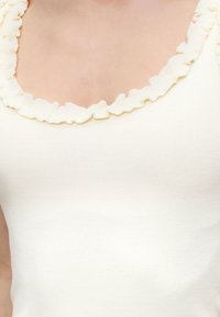 Κοντινό πλάνο ενός αμάνικου τοπ κρεμ χρώματος με στρογγυλή λαιμόκοψη με βολάν στο πάνω μέρος του κορμού ενός ατόμου.