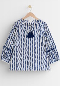 Modrá a bílá blůza s vzorem ve zigzagovém designu, s dlouhými rukávy se stahovacími manžetami a třásní na výstřihu.