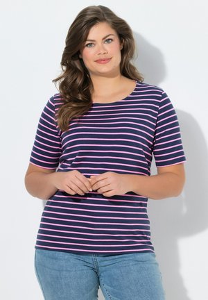 Vrouw met golvend bruin haar die een navyblauw en roze gestreept shirt met korte mouwen en een blauwe spijkerbroek draagt, staat tegen een witte muur.